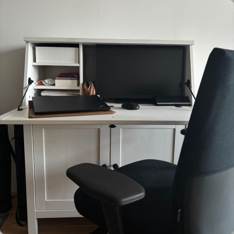 Weißer Schreibtisch mit Monitor, Laptop, Ablagen für diverses Büromaterial und schwarzem Bürostuhl.