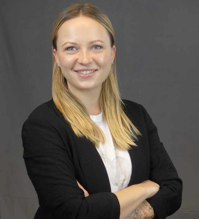 Blonde Frau in schwarzer Jacke lächelt mit verschränkten Armen vor grauem Hintergrund.