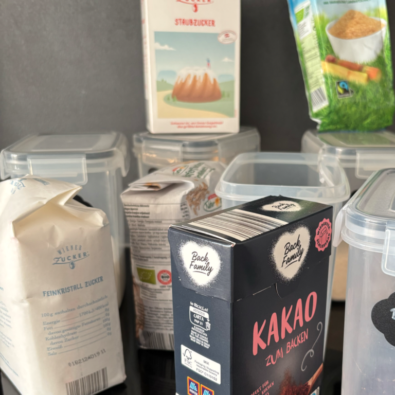 Diverse Lebensmittelverpackungen, darunter Kakao, Mehl und Reis, auf einer Ablage.